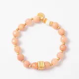 Pulsera elástica compuesta por cuentas de piedra natural en tono rosado, intercaladas con pequeñas esferas doradas. Presenta un detalle central de tres anillos dorados y un dije colgante con forma de moneda con la inscripción MMA.