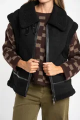 Chaleco estilo gamulán de color negro, con interior y detalles de corderito sintético. Presenta cierre frontal, cuello solapa y bolsillos laterales.