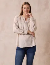 Blusa blanca de lino y rayón, con cuello a la base, escote en V y abertura con botones. Tiene bolsillos en la parte delantera y mangas largas regulables con presilla y botón.