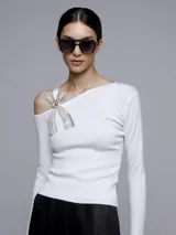 Sweater blanco de punto con escote asimétrico y manga larga. Presenta un broche con forma de moño con apliques de strass en el hombro.