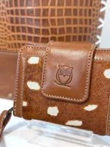 Mini billetera de cuero color canela con aplique de pelo y diseño estampado tipo animal print de bambi con manchas blancas. Cuenta con solapa frontal con cierre de broche y grabado de búho, además de una tira de muñeca desmontable.