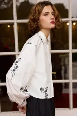 Camisa de algodón color blanco con cuello mao y cierre frontal de botones. Presenta un diseño de bordados florales en color negro distribuidos en el frente y las mangas. Posee mangas largas abullonadas con puños abotonados y un corte recto ligeramente cropped.
