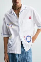 Camisa blanca de algodón con cuello bowling y manga corta, corte relajado. Presenta bordados a contraste en azul, rojo y amarillo que simulan elementos de cocina y artesanía, además de ribetes de pespunte azul en cuello, puños y abertura frontal.