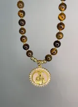Collar de cuentas de piedra natural ojo de tigre marrón con detalles dorados y un dije circular con baño de oro, incrustaciones de circonias y una figura en relieve sobre fondo de nácar.