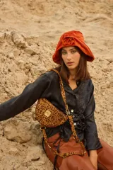Bandolera con silueta de media luna y estructura rígida, confeccionada en cuero con estampado animal print tipo vaca. Presenta herrajes metálicos en tono bronce, cierre superior y correa regulable que permite su uso al hombro o cruzada. Incluye una placa metálica decorativa con iniciales en el frente.