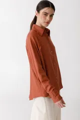 Camisa color ladrillo con rayas verticales finas color crema, de corte recto y mangas largas con puños abotonados. Presenta cuello camisero, cierre frontal con botones y detalle de flecos en los hombros.