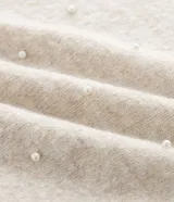 Suéter de punto color beige con cuello redondo, manga larga y aplicaciones de perlas distribuidas por toda la prenda. Presenta puños y ruedo acanalados.