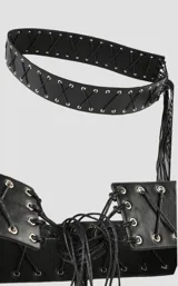 Cinto tipo faja de cuero sintético negro, con diseño de cordones cruzados y ojalillos metálicos plateados. Presenta un extremo con flecos largos decorativos.