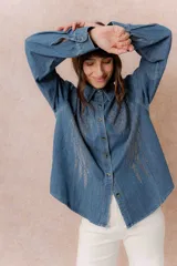 Camisa de denim liviano con calce relajado, cuello camisero clásico y cierre frontal con botones. Presenta una destacada aplicación de strass en el frente formando un diseño de alas.