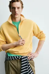 Sudadera de corte relajado confeccionada en algodón con interior de rizo, color amarillo mostaza con efecto lavado. Presenta cuello subido con cierre frontal de cremallera y detalle de cinta trenzada en el tirador, manga larga y acabados en rib.