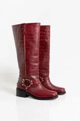 Botas de caña alta en cuero con textura de cocodrilo, color bordó. Presentan un diseño de punta cuadrada, taco bajo y una correa decorativa con argolla metálica y remaches en la zona del tobillo.