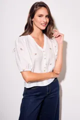 Blusa de viscosa color marrón, con escote en V, mangas cortas abullonadas y cierre frontal con botones. Cuenta con pequeños detalles bordados distribuidos por la prenda.