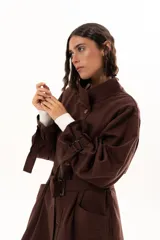 Trench color chocolate con cuello alto, cierre frontal con botones y cinturón ajustable en la cintura. Presenta mangas largas con puños ajustables mediante tiras y hebillas, y bolsillos frontales.