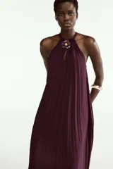 Vestido midi plisado color bordó, con cuello halter, broche de flor en el delantero y anudado en la espalda. Cuenta con forro interior.