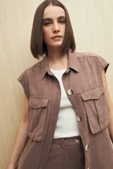 Chaleco color beige con cuello camisero, sin mangas, cierre frontal con botones y cinturón de lazo. Presenta bolsillos de parche en el pecho y pinzas verticales en el frente.