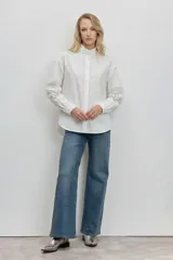 Camisa blanca de corte holgado con cuello alto tipo gorguera y detalles de encaje calado en las mangas. Presenta cierre frontal oculto y puños clásicos.