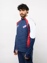 Buzo de entrenamiento Umbro del Club Nacional de Football, con cierre frontal parcial y cuello alto. Presenta un diseño con bloques de color azul, blanco y rojo, con panel frontal acolchado y mangas largas con aberturas para pulgares.