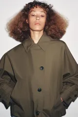 Trench corto color verde militar, con cuello solapa y capucha. Tiene manga larga con trabillas y botón en los puños, bolsillos delanteros y bajo asimétrico. Cierre frontal con botones. El tejido es repelente al agua.