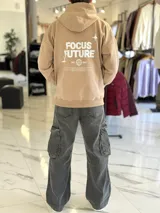 Canguro oversize tostado con capucha y bolsillo tipo canguro, confeccionado con diseño holgado. En la espalda presenta un estampado en color blanco con las palabras "FOCUS FUTURE", gráficos de estrellas y un diseño de globo terráqueo acompañado del texto "EST. 2021".