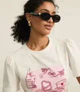 Remera blanca de algodón con cuello redondo y mangas cortas abullonadas. Presenta un estampado con dibujos de objetos femeninos en tonos rosados.