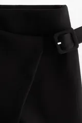 Short pollera negro de tiro alto con diseño cruzado tipo pareo en el frente, asegurado con un cinturón cosido y hebilla revestida. Presenta cierre oculto y cierre de corchete en un lateral.