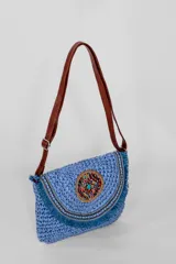 Bandolera de tejido trenzado en color azul, con solapa decorada con flecos y un aplique central circular con cuentas multicolores. Incluye una correa larga ajustable de cuero sintético marrón.