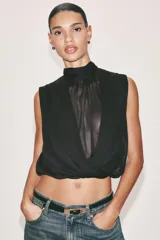 Blusa sin mangas blanca con estampado de lunares negros, cuello alto drapeado y nudo frontal en el bajo.