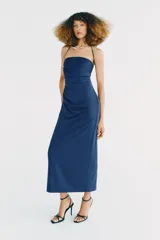 Vestido midi de color azul, con escote recto y cuello halter que incorpora una aplicación metálica tipo choker. Presenta un diseño con frunces laterales en el frente.