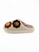 Pantuflas de estar por casa tipo mule, confeccionadas en tejido de peluche color crema. Presentan un diseño con apliques bordados de caritas sonrientes en tonos marrones, bordó y lila. Cuentan con una suela plana de goma y forro interior suave.