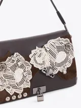 Cartera de hombro color marrón, confeccionada en cuero vacuno con detalles en descarne. Presenta un diseño con apliques de encaje, tachas metálicas y un cierre frontal tipo candado. Posee una estructura semirrígida y correa de cuero.