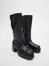 Bota bucanera de cuero vacuno wax color negro, con punta cuadrada, suela de goma con plataforma dentada y taco de madera. Presenta detalle de doble hebilla metálica en la parte superior de la caña.