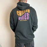 Canguro negro con capucha, de corte regular, con estampado gráfico en la espalda que incluye el texto 'CHILLED VIBES' en colores naranja y violeta, junto con la inscripción 'SANTA BARBARA'.