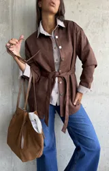 Sobrecamisa de gamuza color chocolate, con cierre frontal de botones metálicos y lazo ajustable en la cintura. Presenta cuello clásico y mangas largas con puños rectos.