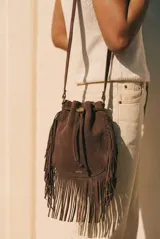 Cartera tipo bucket bag color camel, confeccionada en material sintético con acabado gamuzado. Presenta un diseño decorado con flecos largos en todo el contorno, cierre mediante cordón ajustable y correa larga regulable.