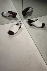 Zueco de piel color blanco, con tacón de madera y plataforma, detalle de tachas metálicas doradas en los laterales y punta cuadrada.