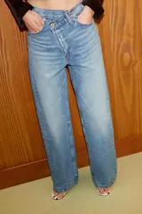 Jeans de tiro alto, color gris oscuro lavado, con pierna ancha extra larga (full length) y diseño de cintura cruzada asimétrica con cierre frontal de cremallera y botones metálicos.