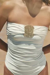 Bañador enterizo tipo bandeau color crema, con escote recto drapeado y detalle de pieza metálica circular dorada en el centro del pecho. Cuenta con forro interior.