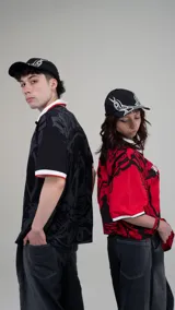 Camisa tipo polo roja de manga corta con cuello blanco y ribetes blancos en las mangas. Presenta un estampado grande en negro con una figura femenina en el centro, la palabra "BLACHAIR" y otros gráficos de estilo gótico/urbano.
