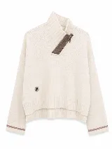 Polera tejida en lana lambswool, de diseño holgado con cuello alto y cierre frontal parcial. Presenta un detalle de parche circular en el frente y rayas en contraste en los puños.