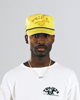 Gorra tipo trucker color amarillo con bordado frontal que incluye la frase 'TWOJEYS' y la silueta de un caballo. Presenta un cordón decorativo negro en la base de la visera y el texto 'CITY COWBOYS' bordado en el lateral.