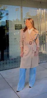 Trench coat color beige de corte clásico, con solapas amplias, doble botonadura frontal, cinturón ajustable con hebilla y trabillas en los puños.