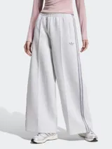 Pantalón deportivo Adidas modelo Spacer, color gris melange, de corte holgado y pierna ancha con pliegues frontales. Cuenta con cintura elástica, bolsillos laterales y las clásicas tiras laterales de la marca en tono gris oscuro, además del logo del Trifolio bordado.
