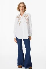 Camisa blanca de corte clásico con cuello camisero y cierre frontal de botones. Presenta un bordado lineal en color marrón oscuro en ambos lados del frente, representando motivos florales.
