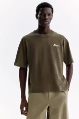 Remera de algodón verde militar con efecto lavado, cuello redondo de canalé, mangas cortas con hombros caídos y pequeño estampado tipográfico en el pecho.