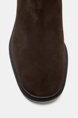Bota de caña alta estilo hípico confeccionada en serraje color marrón oscuro. Presenta punta redonda, suela plana con pequeño tacón y detalle de tira con hebilla metálica en la parte superior de la caña.
