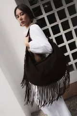 Bolso tipo hobo de gran tamaño confeccionado en gamuza color marrón oscuro. Presenta un diseño de media luna con flecos largos en todo el borde inferior y lateral, y un asa ancha con detalles de pespuntes artesanales.