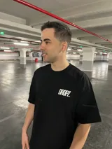 Remera de manga corta color beige natural, corte relajado y amplio. Presenta un estampado tipográfico grande en la espalda con el texto "HOP ON THE RIDE" en negro, y debajo el logo "DROPZ STUDIO EST. 2025".