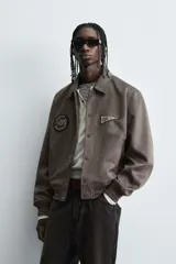 Cazadora estilo varsity de tejido efecto ante en color beige. Presenta cuello solapa, cierre frontal con botones a presión y puños y ruedo con acabado en rib a rayas negras. Incluye parches bordados decorativos en el pecho y la manga, además de bolsillos laterales de doble vivo.
