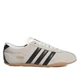 Championes Adidas modelo Tokyo de corte bajo, confeccionados en gamuza color beige con las icónicas tres tiras laterales en cuero negro. Presenta suela de caucho color caramelo y detalle de logo dorado en el lateral.