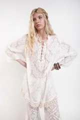 Vestido camisero corto de encaje blanco con diseño ornamental, escote en V con cordón ajustable y ojales metálicos, mangas 3/4 acampanadas y ruedo con detalle de flecos de caracoles tejidos a mano.